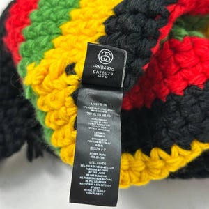 May include: A colorful, hand-crocheted beanie in a Rastafarian color scheme of red, yellow, green, and black. The beanie features a tag with the brand logo and care instructions. The L/XL/G/TG size is indicated on the tag.