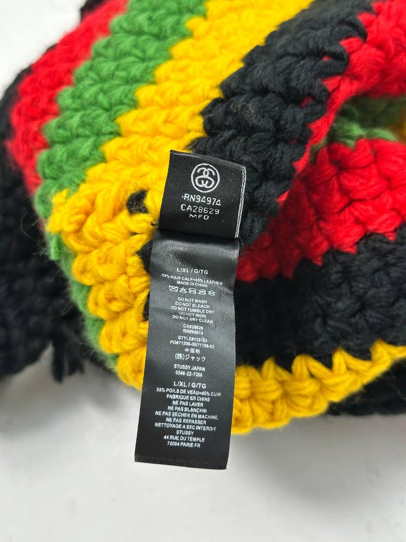 May include: A colorful, hand-crocheted beanie in a Rastafarian color scheme of red, yellow, green, and black. The beanie features a tag with the brand logo and care instructions. The L/XL/G/TG size is indicated on the tag.