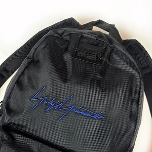 Peut inclure: Un sac &agrave; dos noir en tissu synth&eacute;tique textur&eacute; avec une signature brod&eacute;e en bleu sur la poche avant. Le sac comprend une boucle de fermeture en m&eacute;tal noir sur le dessus et des bretelles rembourr&eacute;es pour le transport.