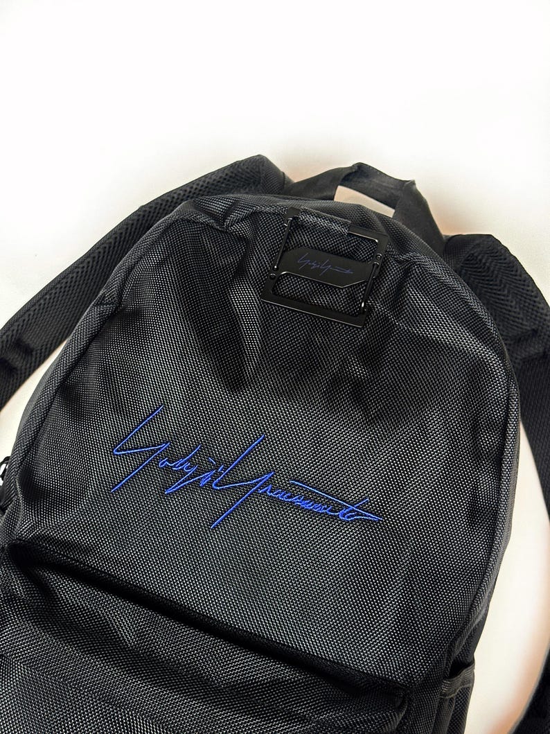 Peut inclure: Un sac &agrave; dos noir en tissu synth&eacute;tique textur&eacute; avec une signature brod&eacute;e en bleu sur la poche avant. Le sac comprend une boucle de fermeture en m&eacute;tal noir sur le dessus et des bretelles rembourr&eacute;es pour le transport.