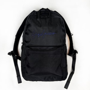 Peut inclure: Un sac &agrave; dos noir en tissu textur&eacute;, dot&eacute; d'un grand compartiment principal et d'une poche avant plus petite. L'avant pr&eacute;sente une signature brod&eacute;e en bleu. Le sac comprend des bretelles r&eacute;glables et une poign&eacute;e de transport sup&eacute;rieure.