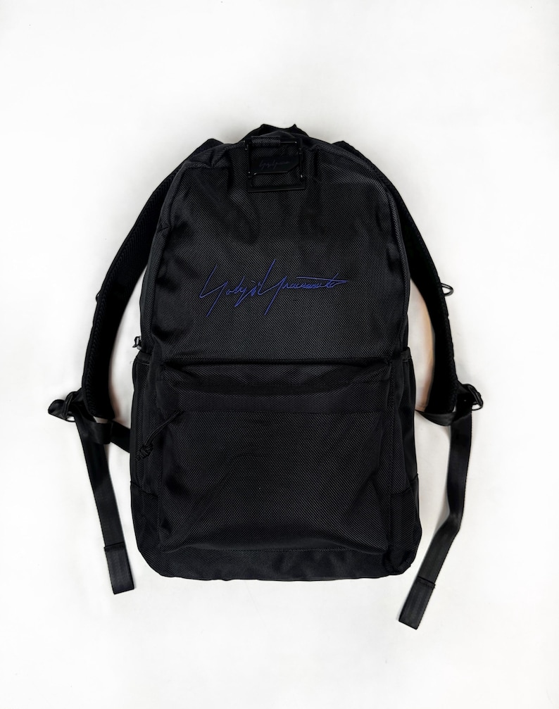 Peut inclure: Un sac &agrave; dos noir en tissu textur&eacute;, dot&eacute; d'un grand compartiment principal et d'une poche avant plus petite. L'avant pr&eacute;sente une signature brod&eacute;e en bleu. Le sac comprend des bretelles r&eacute;glables et une poign&eacute;e de transport sup&eacute;rieure.