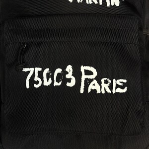 Puede incluir: Una mochila negra con texto blanco de estilo pintado a mano. La parte superior muestra las palabras SAINT MARTIN, mientras que el bolsillo frontal presenta el texto 75003 PARIS. La bolsa est&aacute; hecha de un material de tela duradero y texturizado.