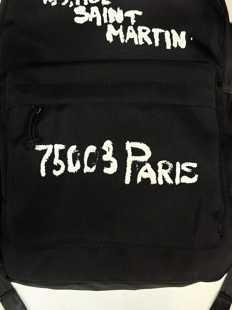 Puede incluir: Una mochila negra con texto blanco de estilo pintado a mano. La parte superior muestra las palabras SAINT MARTIN, mientras que el bolsillo frontal presenta el texto 75003 PARIS. La bolsa est&aacute; hecha de un material de tela duradero y texturizado.