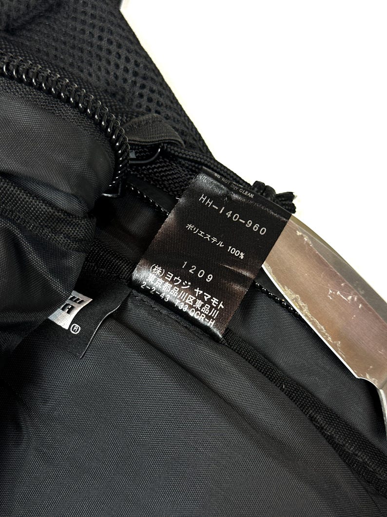 Peut inclure: Un sac en textile noir dot&eacute; d'une &eacute;tiquette d'entretien int&eacute;rieure bien visible avec le texte HH-140-960, 100% polyester, et des caract&egrave;res japonais incluant Yohji Yamamoto. Le sac pr&eacute;sente une section en maille noire, une fermeture &eacute;clair et une doublure en tissu noir lisse.