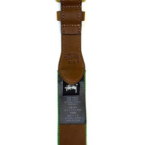 Puede incluir: Un cintur&oacute;n de cuero marr&oacute;n con una hebilla de metal color lat&oacute;n. La correa de cuero presenta el logotipo de St&uuml;ssy grabado cerca de la hebilla y una etiqueta de tela negra con texto blanco que incluye CA# 28629, RN# 94974, STYLE# 335003 y composici&oacute;n.