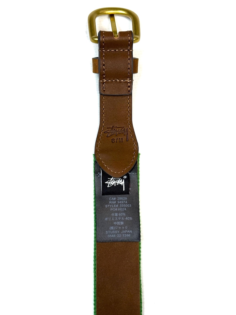 Puede incluir: Un cintur&oacute;n de cuero marr&oacute;n con una hebilla de metal color lat&oacute;n. La correa de cuero presenta el logotipo de St&uuml;ssy grabado cerca de la hebilla y una etiqueta de tela negra con texto blanco que incluye CA# 28629, RN# 94974, STYLE# 335003 y composici&oacute;n.