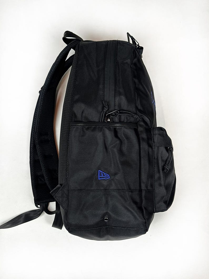 Peut inclure: Un sac &agrave; dos noir dot&eacute; d'un ext&eacute;rieur en tissu textur&eacute;, d'une poche lat&eacute;rale avec un petit logo brod&eacute; bleu et de bretelles rembourr&eacute;es r&eacute;glables. Le design comprend un compartiment avant zipp&eacute; et des tirettes de fermeture &eacute;clair noires.