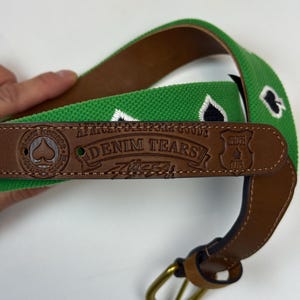 Puede incluir: Un cintur&oacute;n tejido verde con s&iacute;mbolos de picas negros bordados. El cintur&oacute;n incluye una punta de cuero marr&oacute;n grabada con el texto African Diaspora Goods, Denim Tears y Spades Club, junto con una hebilla de metal color lat&oacute;n y una trabilla.
