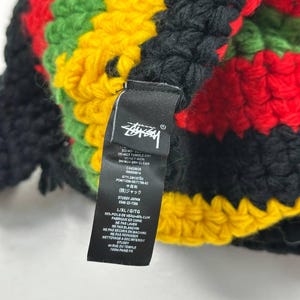 May include: A multi-colored crochet hat in black, red, green, and yellow. The hat features a tag with the Stussy logo and care instructions. The hat is made of a soft, textured material.
