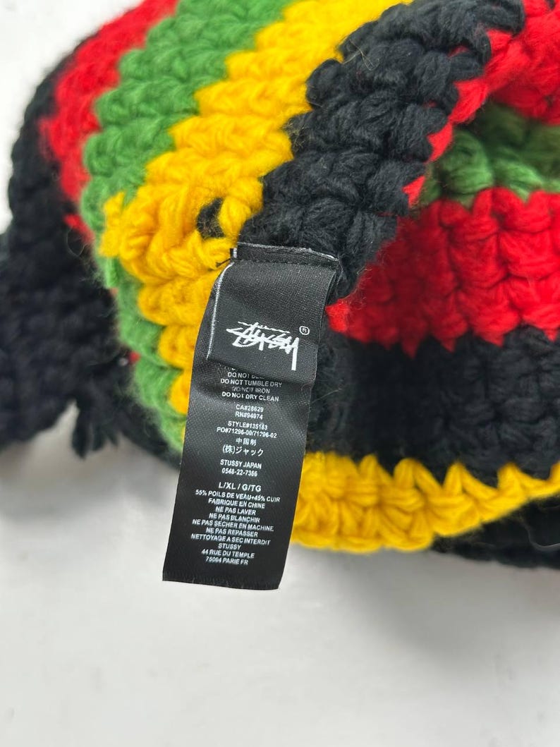 May include: A multi-colored crochet hat in black, red, green, and yellow. The hat features a tag with the Stussy logo and care instructions. The hat is made of a soft, textured material.