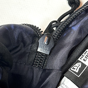 Puede incluir: Un bolso de tela con estampado azul oscuro y negro que presenta un tirador de cremallera YKK negro y una extensi&oacute;n de cord&oacute;n negro. Se ve una etiqueta interior blanca con el logo de New Era, 100% poli&eacute;ster y fabricado en Vietnam.