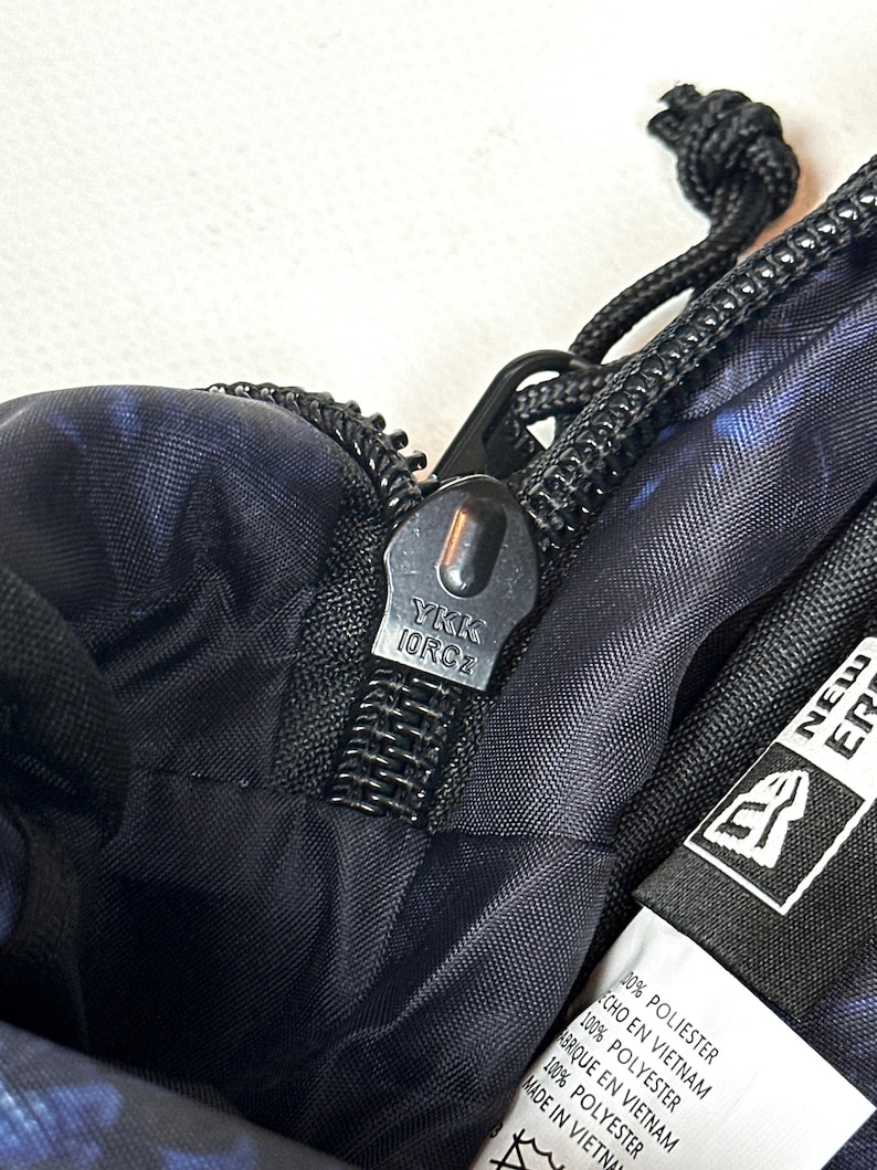 Puede incluir: Un bolso de tela con estampado azul oscuro y negro que presenta un tirador de cremallera YKK negro y una extensi&oacute;n de cord&oacute;n negro. Se ve una etiqueta interior blanca con el logo de New Era, 100% poli&eacute;ster y fabricado en Vietnam.
