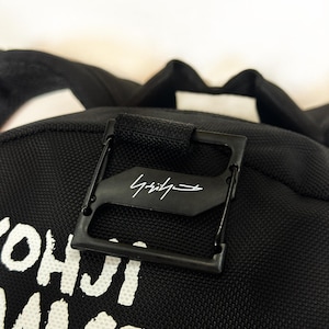 Puede incluir: Una mochila negra con una superficie de tela texturizada y texto impreso en blanco que dice YOHJI YAMAMOTO. Una pieza de metal cuadrada de color negro est&aacute; sujeta en la parte superior, mostrando un logotipo de firma blanco en el centro.