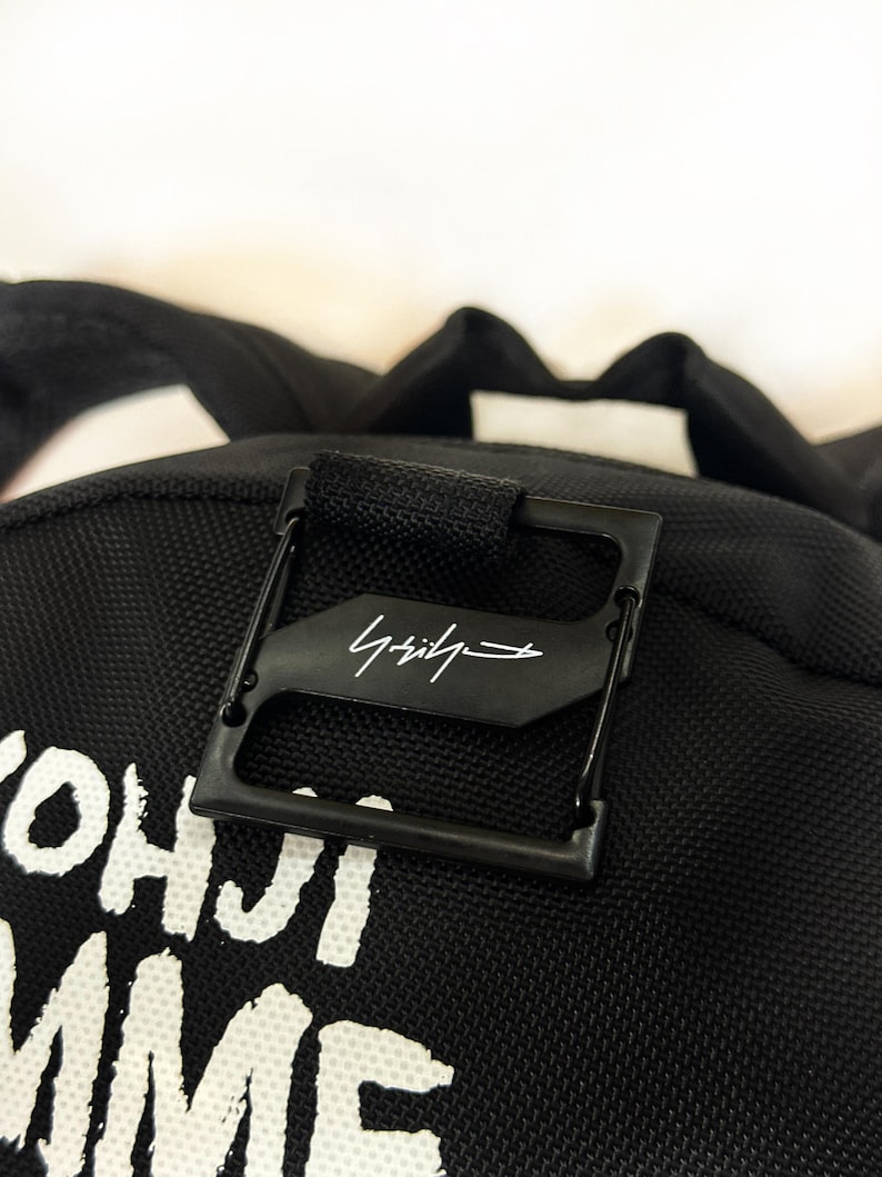 Puede incluir: Una mochila negra con una superficie de tela texturizada y texto impreso en blanco que dice YOHJI YAMAMOTO. Una pieza de metal cuadrada de color negro est&aacute; sujeta en la parte superior, mostrando un logotipo de firma blanco en el centro.
