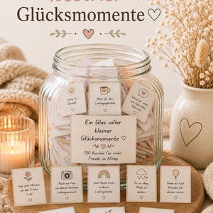 May include: A clear glass jar filled with small square cards, each featuring a unique illustration and German text. A label on the jar reads 'Ein Glas voller kleiner Glücksmomente' with '150 Karten für mehr Freude im Alltag'. Several cards are displayed in front.