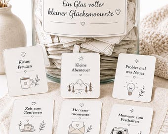 Puede incluir: Un frasco de vidrio transparente con tapa de corcho lleno de pequeñas tarjetas blancas, cada una con texto negro e ilustraciones de líneas. La etiqueta del frasco dice Ein Glas voller kleiner Glücksmomente. Varias tarjetas se muestran al frente, con frases como Kleine Freuden, Kleine Abenteuer y Probier mal was Neues.
