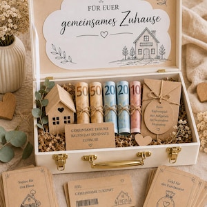 May include: A white wooden gift box containing rolled banknotes, a small wooden house, and brown paper cards with German text. The lid interior features a cloud-shaped sign reading FÜR EUER gemeinsames Zuhause with a house illustration.