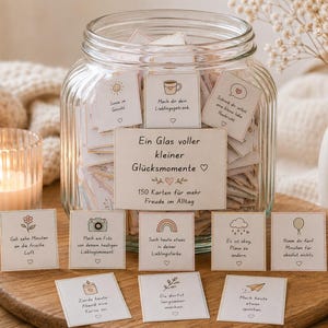 Puede incluir: Un frasco de vidrio transparente lleno de peque&ntilde;as tarjetas de papel, con una etiqueta que dice Ein Glas voller kleiner Gl&uuml;cksmomente, 150 Karten f&uuml;r mehr Freude im Alltag. Varias tarjetas individuales con ilustraciones se disponen delante.