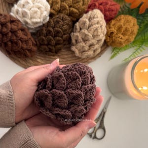 May include: A dark brown crochet pinecone held in two hands, with a collection of various colored crochet pinecones in the background. The text MushyyCrochet is visible on the dark brown pinecone. The scene includes a candle and scissors.