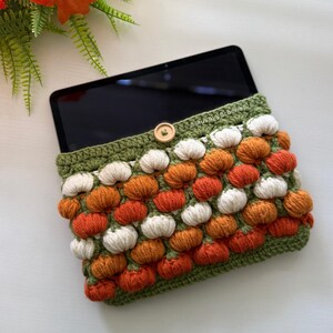 May include: A rectangular crochet tablet sleeve featuring a textured pattern of small white and orange pumpkins on a green base. The sleeve has a green crocheted border, a single light-coloured wooden button closure, and the text MushyyCrochet.