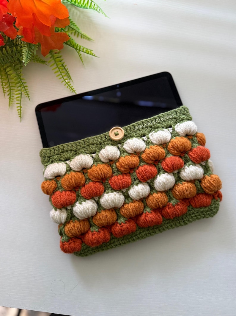 May include: A rectangular crochet tablet sleeve featuring a textured pattern of small white and orange pumpkins on a green base. The sleeve has a green crocheted border, a single light-coloured wooden button closure, and the text MushyyCrochet.