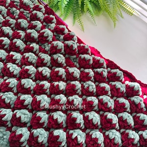 May include: A textured crochet blanket featuring a repeating pattern of raised bobble stitches in deep burgundy and soft sage green yarn. The fabric displays a dense, plush construction with the text MushyyCrochet visible on the surface.