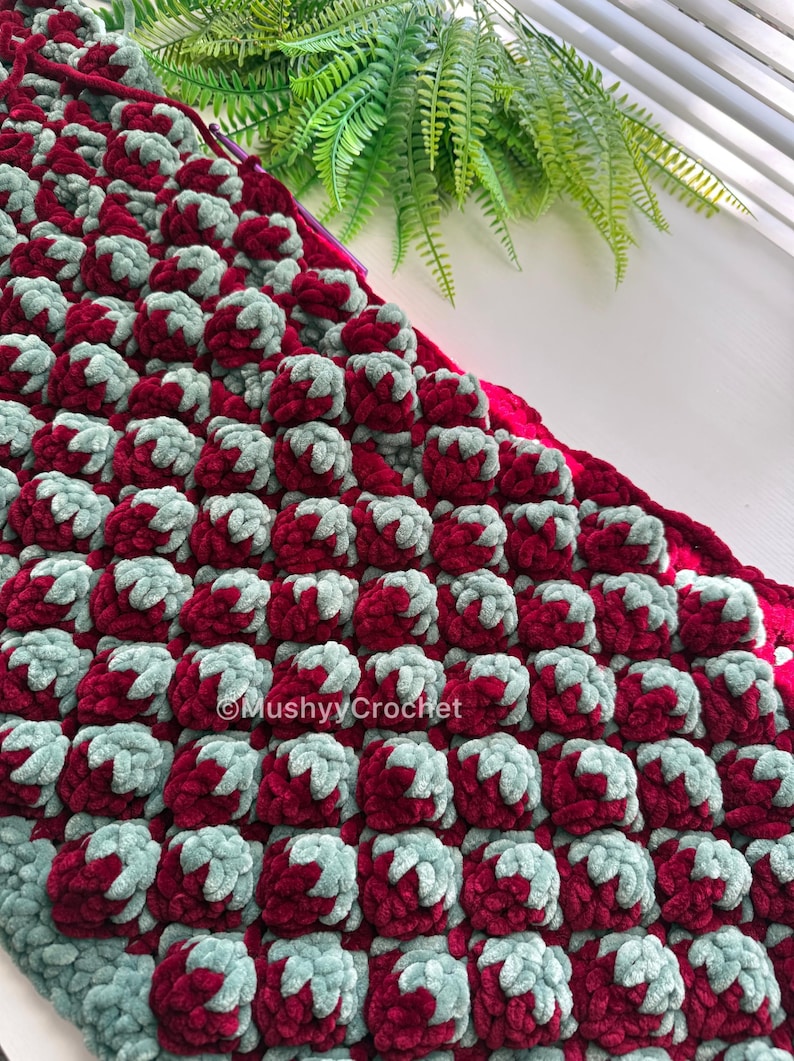 May include: A textured crochet blanket featuring a repeating pattern of raised bobble stitches in deep burgundy and soft sage green yarn. The fabric displays a dense, plush construction with the text MushyyCrochet visible on the surface.