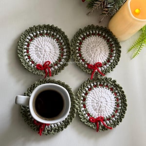 May include: Four round, crocheted coasters featuring a white center, a red inner ring, and a dark green outer border with a small red bow accent. One coaster supports a white ceramic mug filled with dark coffee, while the others are empty.
