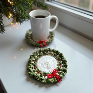 Puede incluir: Dos posavasos redondos de ganchillo con un centro blanco rodeado por un borde verde texturizado que recuerda a una corona, decorados con pequeños lazos de lana roja. Un posavasos está bajo una taza de cerámica blanca y otro está plano delante.
