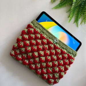 May include: A rectangular crochet tablet sleeve featuring a textured strawberry pattern in red and green yarn. The sleeve has a green crocheted border, a clear button closure, and contains a tablet with a colorful screen display.