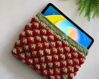May include: A rectangular crochet tablet sleeve featuring a textured strawberry pattern in red and green yarn. The sleeve has a green crocheted border, a clear button closure, and contains a tablet with a colourful screen display.