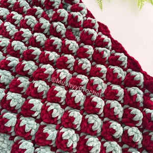 May include: A textured crochet blanket featuring a repeating pattern of raised bobble stitches in alternating deep burgundy and soft sage green yarn. The fabric has a thick, plush appearance with the text MushyyCrochet printed on the surface.