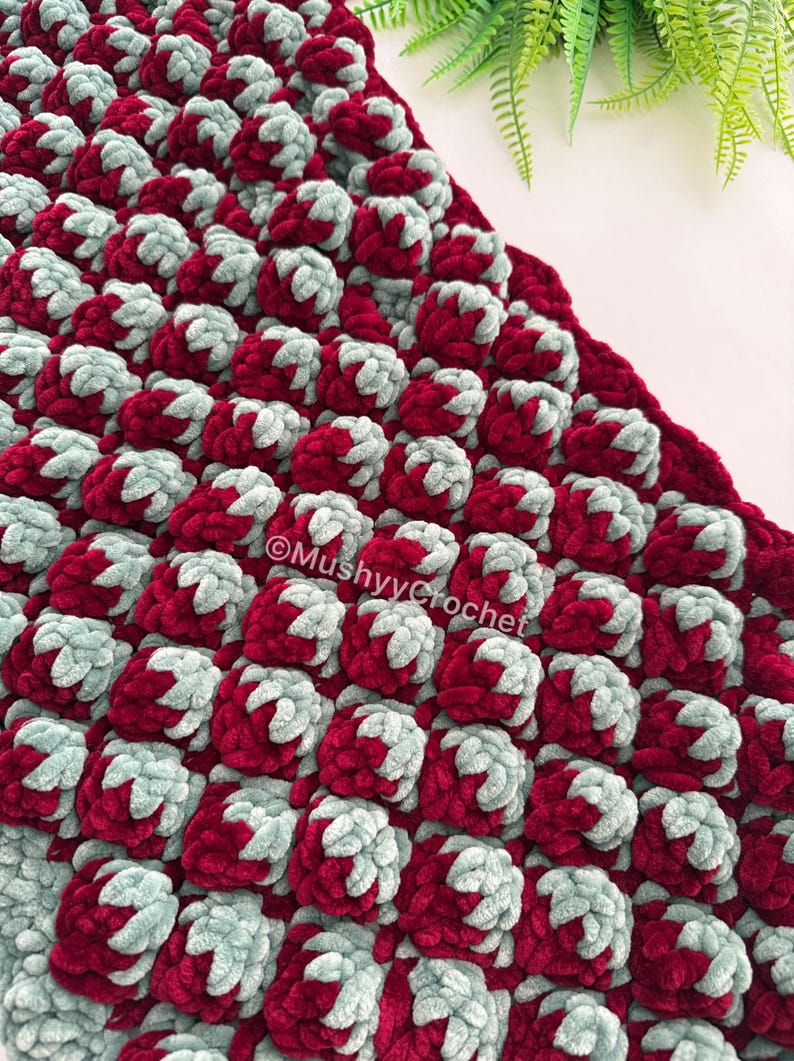 May include: A textured crochet blanket featuring a repeating pattern of raised bobble stitches in alternating deep burgundy and soft sage green yarn. The fabric has a thick, plush appearance with the text MushyyCrochet printed on the surface.