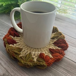 May include: A white ceramic mug sits on a round, beige crocheted coaster featuring a border of textured, leaf-shaped yarn clusters in shades of dark green, rust orange, and brown. The text MushyyCrochet is visible on the coaster surface.