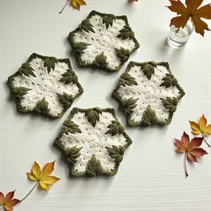 Puede incluir: Cuatro posavasos hexagonales de ganchillo con un centro blanco de patrón estrellado y motivos en forma de hoja de color verde oliva oscuro en los bordes. Cada posavasos presenta una apariencia texturizada y hecha a mano en blanco y verde.