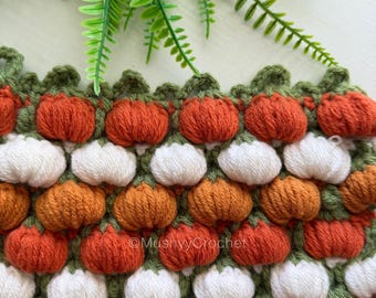 May include: A crocheted fabric featuring a repeating pattern of small, puffed pumpkin shapes in shades of orange, white, and rust, separated by green yarn accents. The text MushyyCrochet is visible in the center of the fabric.