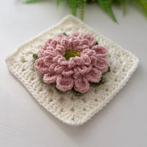 May include: A square crochet granny square in off-white yarn featuring a three-dimensional layered pink flower with a lime green center. The text MushyyCrochet is visible on the petals. The design consists of intricate yarn loops and stitches.