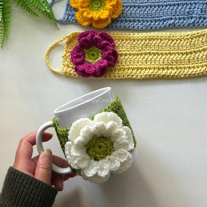 Puede incluir: Una taza de cerámica blanca con una funda de ganchillo verde que presenta una gran flor blanca. Se ven dos tiras de ganchillo adicionales, una azul claro con una flor amarilla y otra amarilla con una flor morada, cada una con un centro verde.