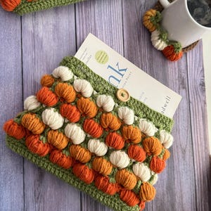 May include: A crochet book sleeve featuring a repeating pattern of small orange and cream-coloured pumpkins on a green background, secured with a single wooden button. A white mug with a matching pumpkin-themed crochet coaster sits nearby.