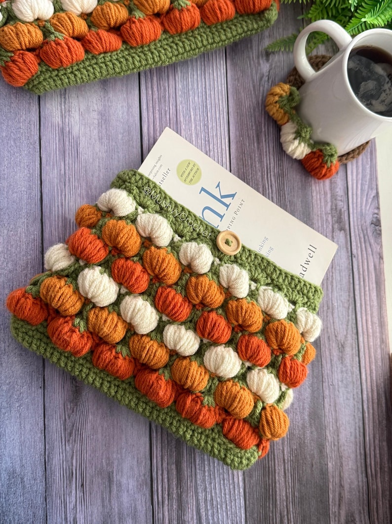 May include: A crochet book sleeve featuring a repeating pattern of small orange and cream-coloured pumpkins on a green background, secured with a single wooden button. A white mug with a matching pumpkin-themed crochet coaster sits nearby.