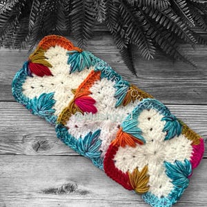 Puede incluir: Tres motivos cuadrados de ganchillo con un centro blanco y detalles en forma de pétalos en tonos verde azulado, naranja, rojo y amarillo mostaza. El texto @MushyCrochet es visible en el centro de los cuadrados, dispuestos en fila.