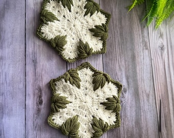 May include: Two hexagonal crochet coasters featuring a cream-colored center with a starburst pattern and dark olive green leaf-shaped clusters along the edges. The text "Mushy" is visible on the lower coaster, which has a textured yarn construction.