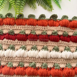May include: A crocheted fabric featuring horizontal rows of textured pumpkin motifs in orange, red, and white, separated by beige yarn bands. Each pumpkin has a small green stem. The text MushyyCrochet is visible in the center of the fabric.