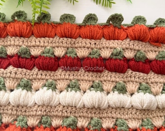 May include: A crocheted fabric featuring horizontal rows of textured pumpkin motifs in orange, red, and white, separated by beige yarn bands. Each pumpkin has a small green stem. The text MushyyCrochet is visible in the center of the fabric.