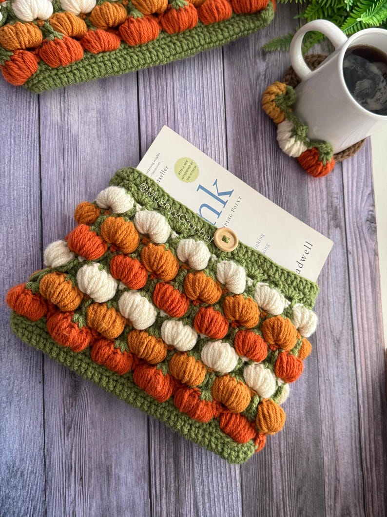 May include: A crochet book sleeve featuring a repeating pattern of small orange and white pumpkins on a green background, secured with a single wooden button. The sleeve holds a book with the visible text Blink and MushyCrochet.