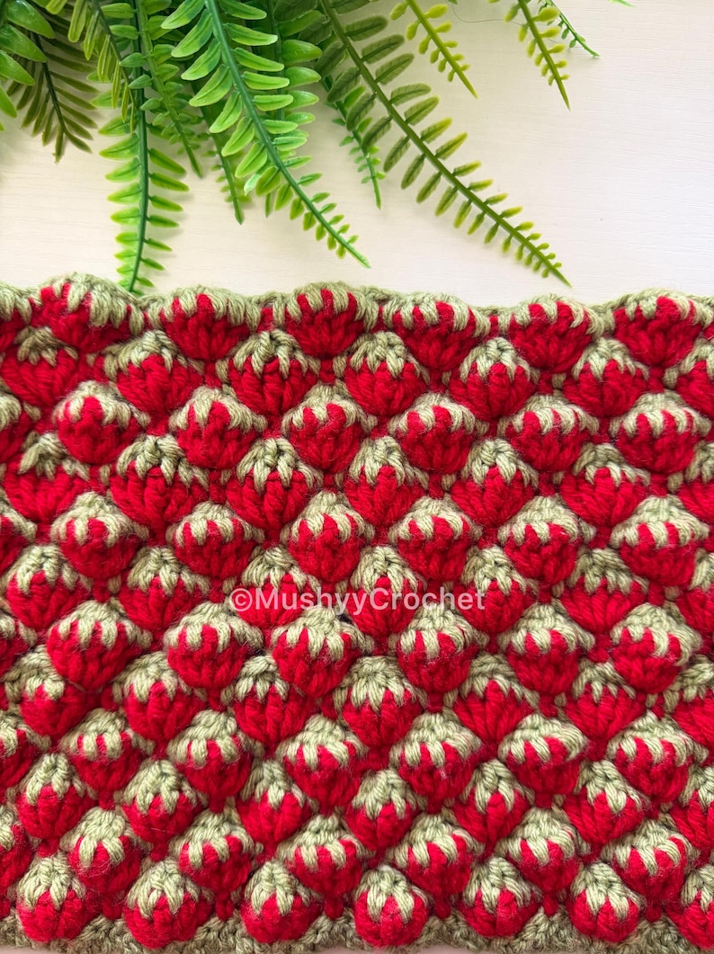 May include: A crocheted fabric featuring a repeating strawberry pattern in bright red and sage green yarn. The texture is raised and bumpy, with each strawberry shape topped by green leaves. The text MushyyCrochet is printed in white in the center.