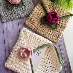 May include: Three square crochet envelopes in grey, tan, and cream, each featuring a green crocheted border and a three-dimensional crocheted flower in pink or dark magenta. The cream envelope is partially open, revealing a white card with text.