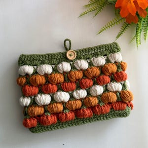 May include: A rectangular crochet pouch featuring a repeating pattern of small white and orange pumpkin-shaped bobble stitches on a dark green background. The pouch has a single wooden button closure at the top and the text MushyyCrochet.