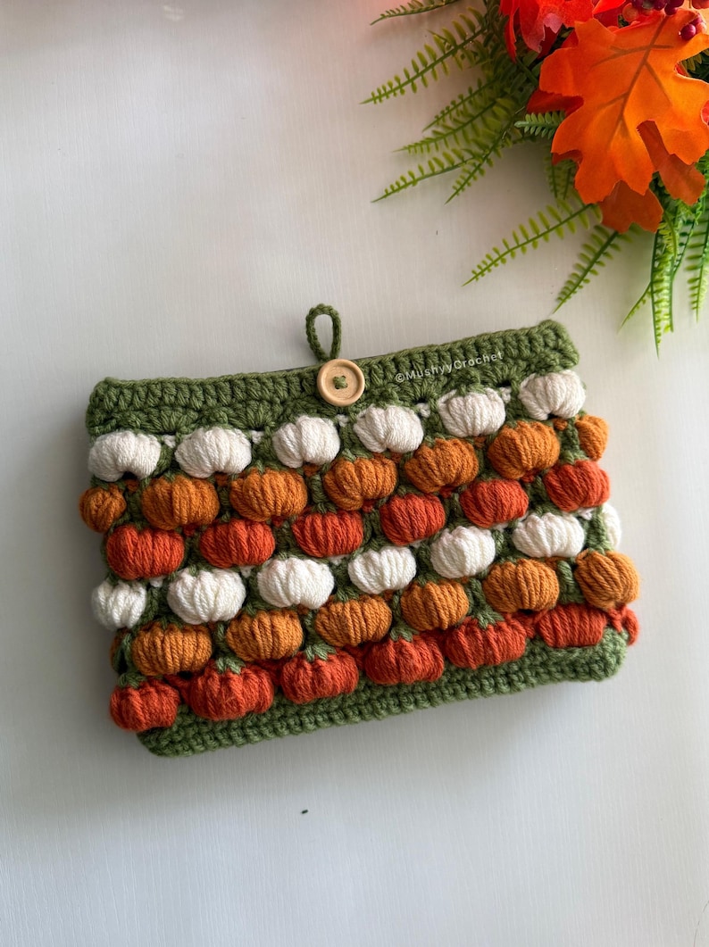 May include: A rectangular crochet pouch featuring a repeating pattern of small white and orange pumpkin-shaped bobble stitches on a dark green background. The pouch has a single wooden button closure at the top and the text MushyyCrochet.